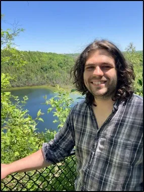 A portrait of Mac Carden. He is smiling in a plaid shirt in front of a lake. 